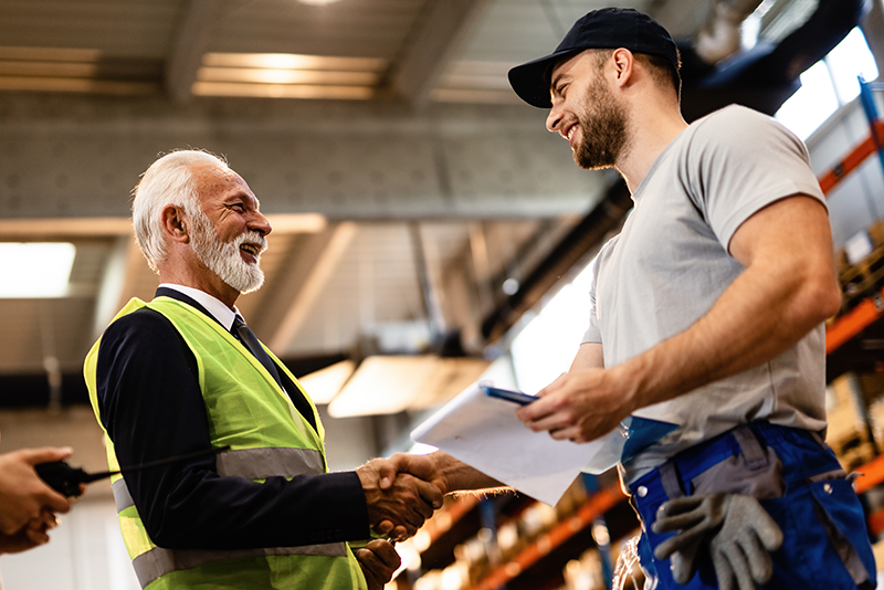 Ein Büromitarbeiter und ein Mitarbeitender aus dem Handwerk schütteln sich lächelnd die Hand